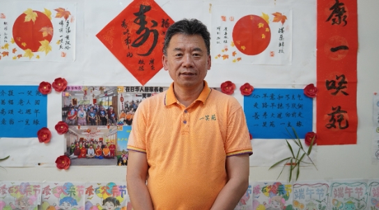介護施設「一笑苑」内の壁に飾られた写真や書画の作品の前に立つ「一笑苑」代表取締役の佐々木弘志さん(撮影・許可)。